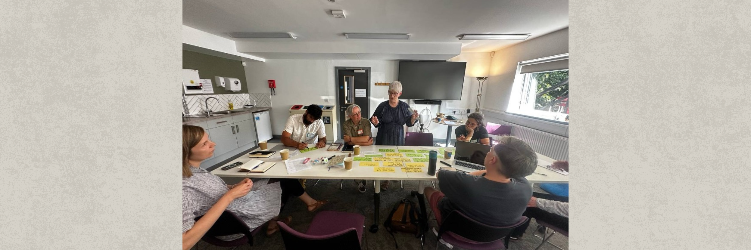 Photograph of a group meeting in a conference room with six people gathered around a table covered in papers and sticky notes, engaged in discussion. A facilitator stands in the middle.  The room features a large TV screen, windows, a white table with post-it notes on for a collaborative brainstorming session.