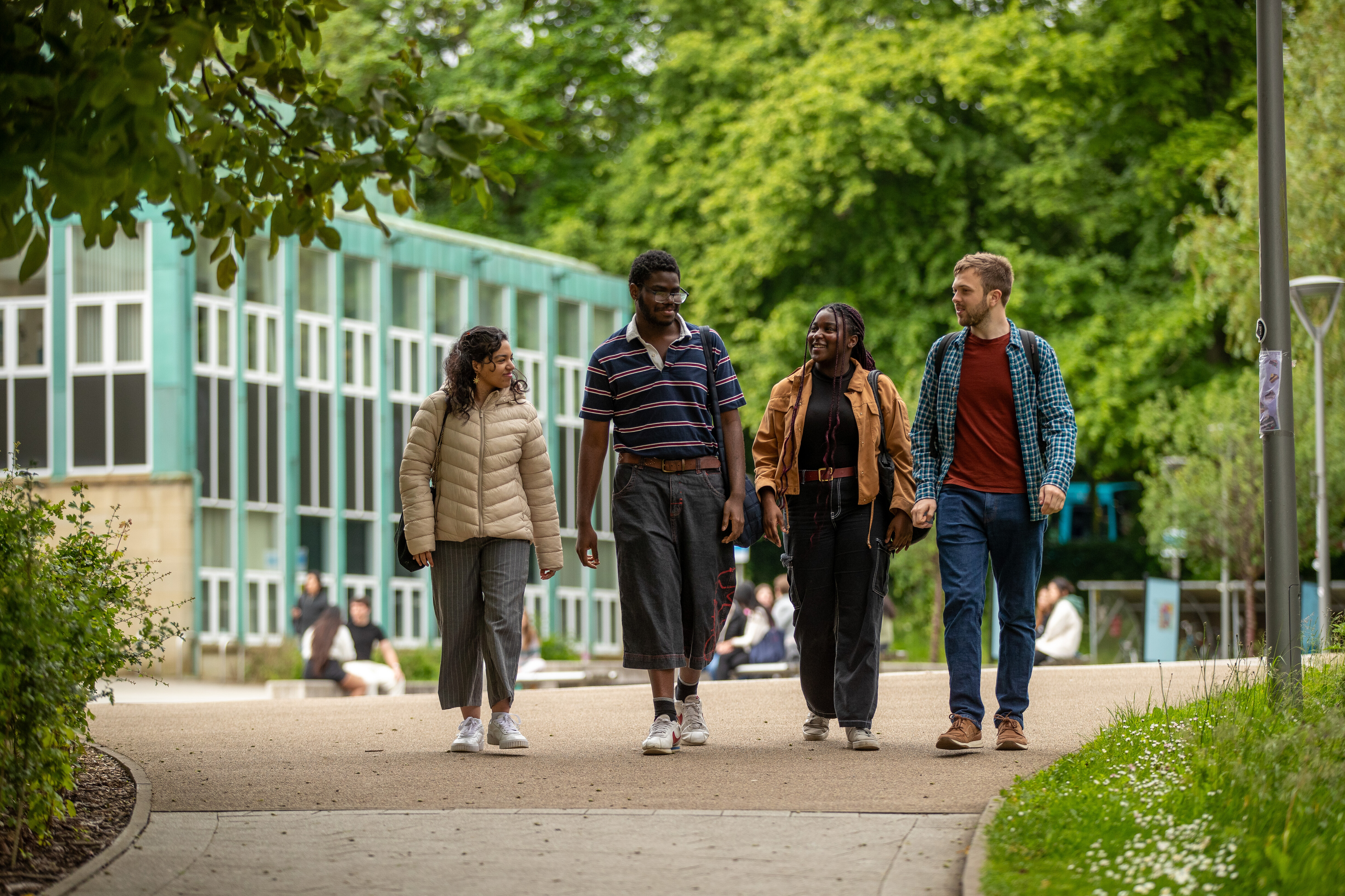 Four students walk towards the viewer. They are chatting as they walk. In the background are a mix of buildings include a red brick building and zinc green building and lots of trees (including a willow tree) in full leaf and grassy banks. It’s an overcast day.