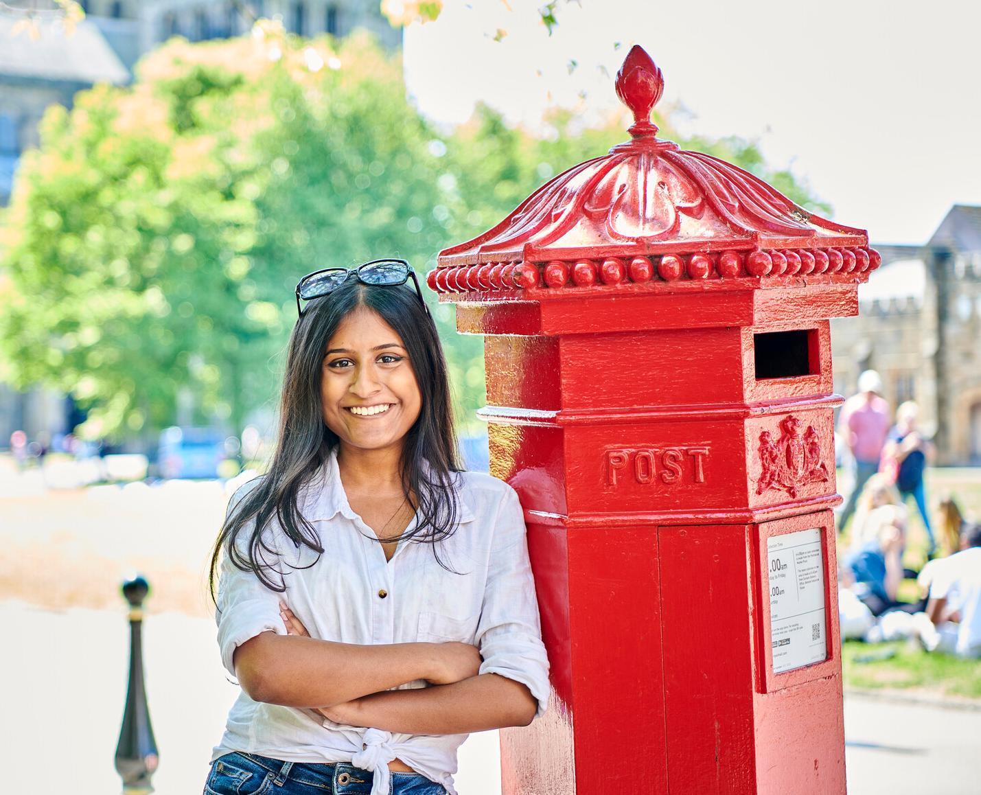 Student leaning on red post box at Palace Green smiling