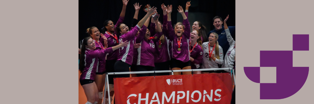 A women's volleyball team standing behind a red 'Champions' banner, holding a trophy high, and celebrating.
