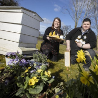 A man and woman smile towards the camera. They are outside, and it's a bright, sunny day. There is a white bee hive to the left of the couple and bright yellow daffodils to the right. They are holding honey jars and honey combs.
