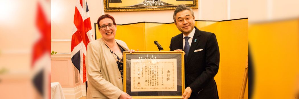 A woman and a man hold a framed certificate and smile at the camera. The certificate is written in Japanese.