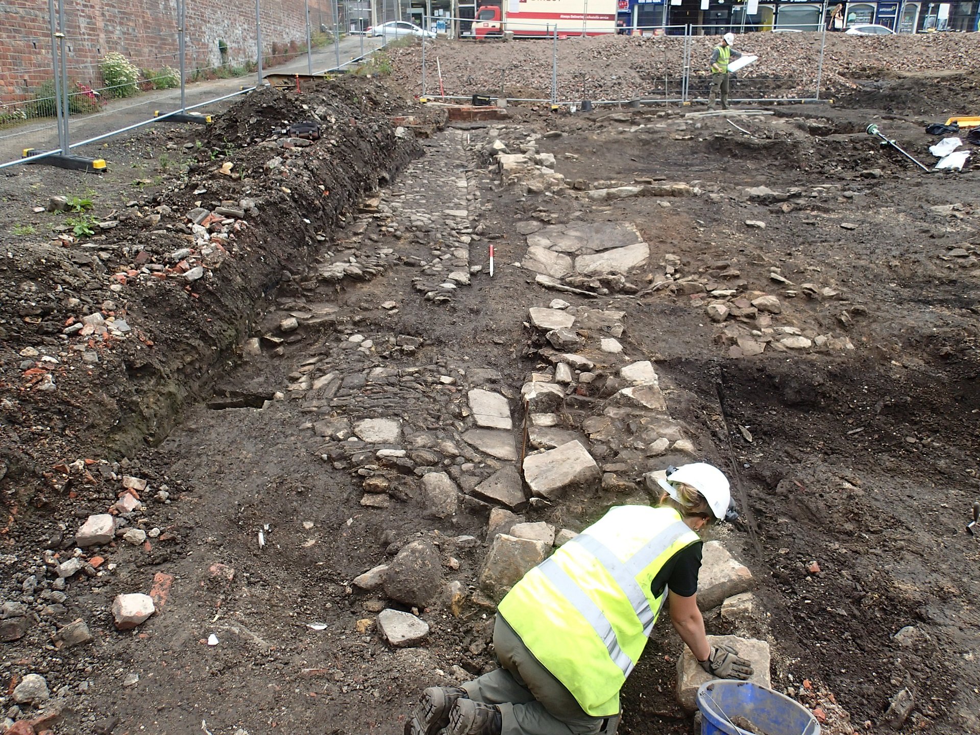 A coloured photograph showing archaeologists on a site in Claypath. The remains of a wall can be seen on the right.