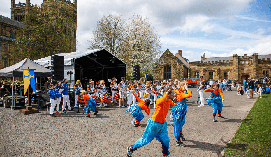 A group of drummers and performers dressed in orange and blue on Palace Green